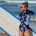 Modelo usando maiô esportivo de manga longa com estampa floral e zíper frontal. Ideal para práticas esportivas, oferece proteção UV e visual sofisticado à beira-mar.
